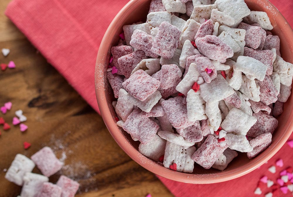 A bowl of pink and white Strawberries & Cream Puppy Chow (Muddy Buddies), dusted with powdered sugar and decorated with heart-shaped sprinkles, placed on a rustic wooden table with a pink napkin. A few pieces of the snack are scattered around, enhancing the festive and sweet atmosphere.
