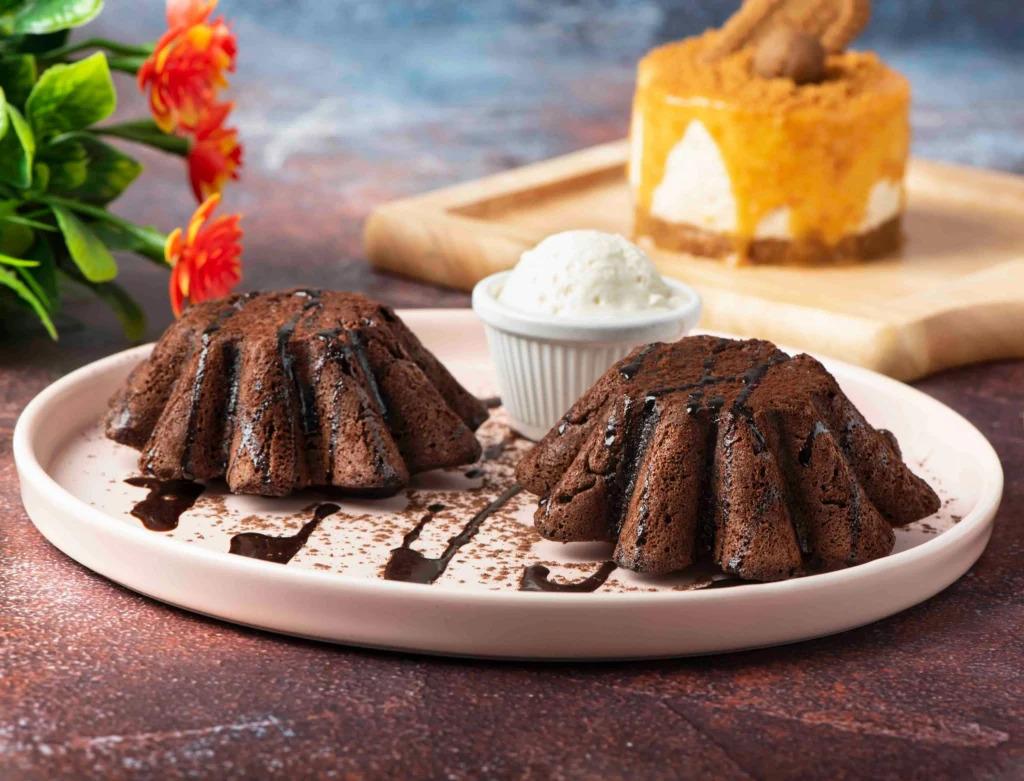 Two rich chocolate lava cakes drizzled with chocolate sauce, served with a scoop of vanilla ice cream, with a caramel cheesecake in the background.